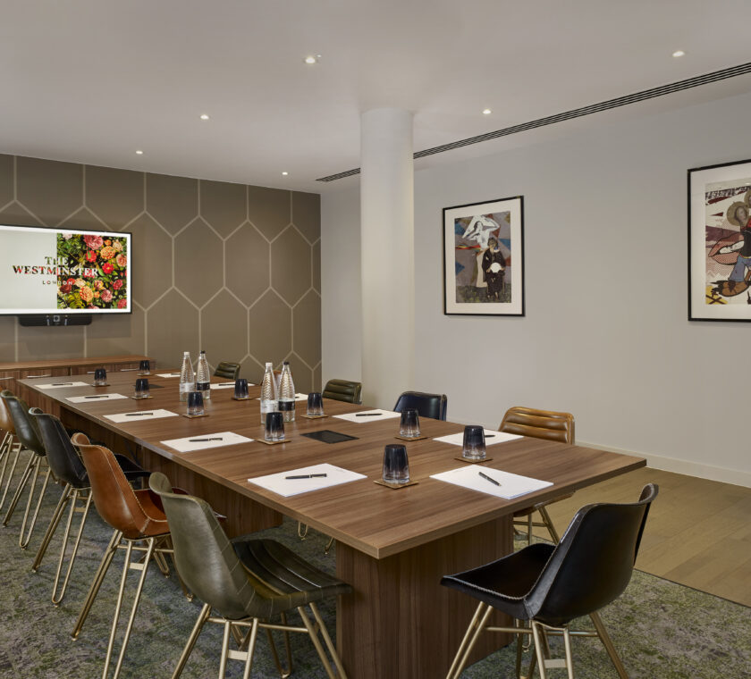 Meeting room set up in boardroom style. Long table with chairs all around. The TV screen has floral TW logo and there are two art pieces on the walls. There are no persons in the photograph.