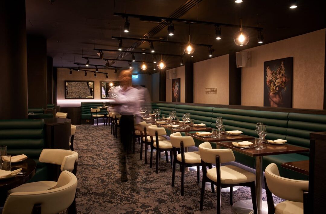 Hero image inside The Westminster Bar & Grill features unidentified person walking pass tables and green benches with white single chairs.